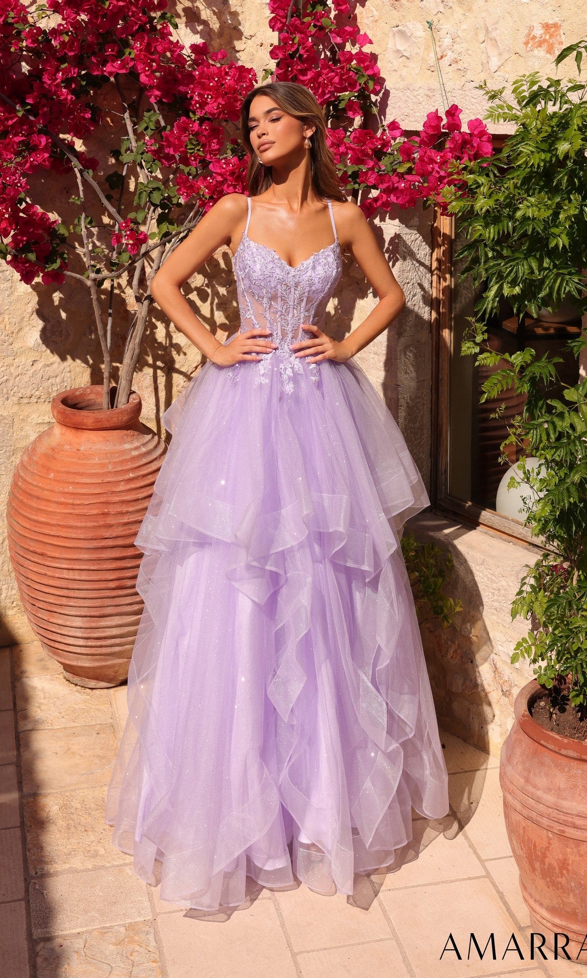 A woman stands outdoors by potted plants, wearing the Amarra 89504 Lace-Up Long Tiered Prom Dress in lilac, featuring thin straps, floral detailing, and a fluffy skirt.